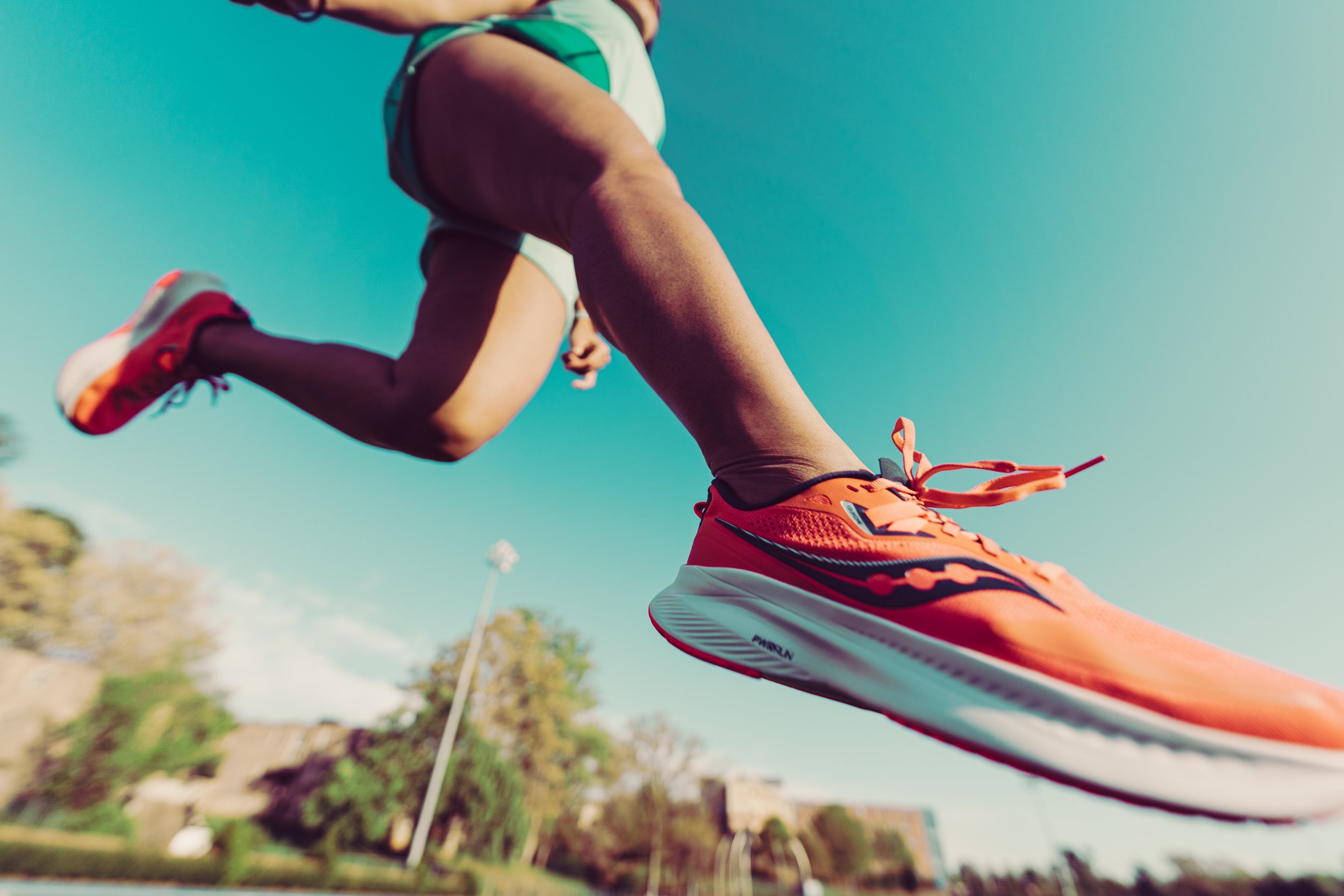 Close-up of an orange running shoe and a person mid-air during a run outdoors on a sunny day.