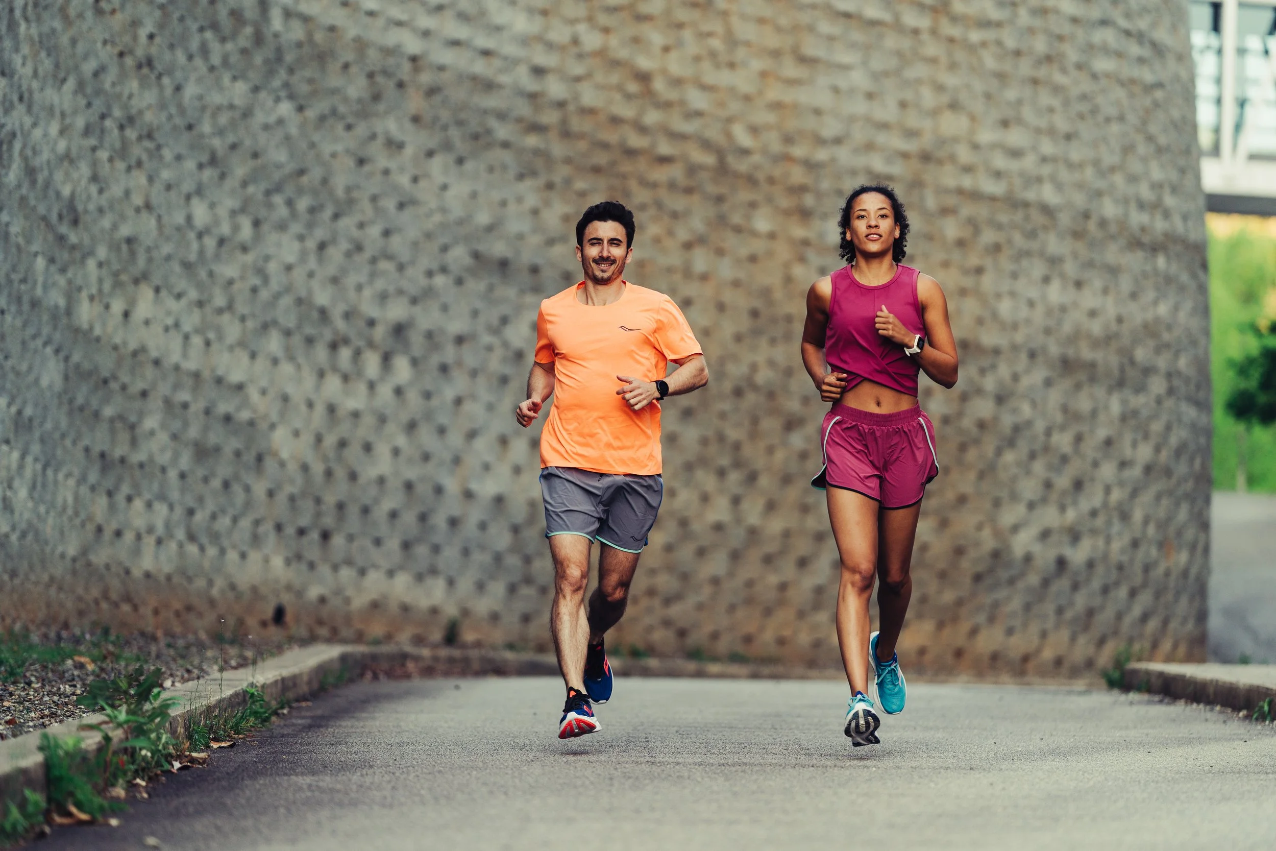 Two people jogging outdoors on a paved path beside a large textured concrete wall.