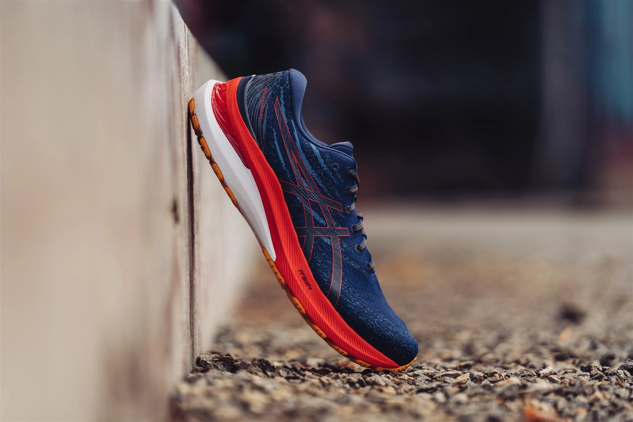 A blue and red running shoe resting against a concrete wall on a gravel surface.