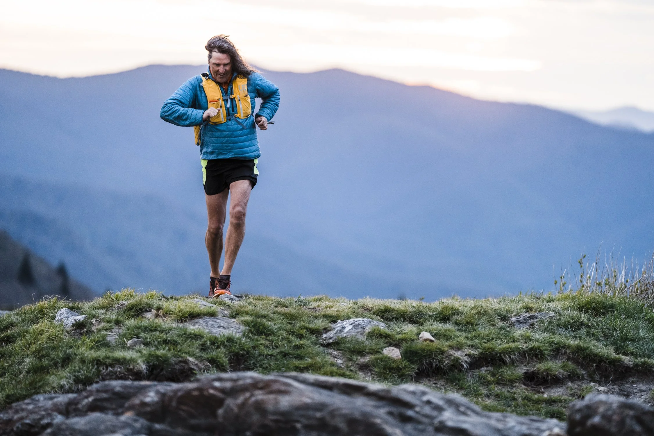 A man in running gear, wearing a blue jacket and black shorts, is running on a grassy trail with mountains in the background, during sunset.