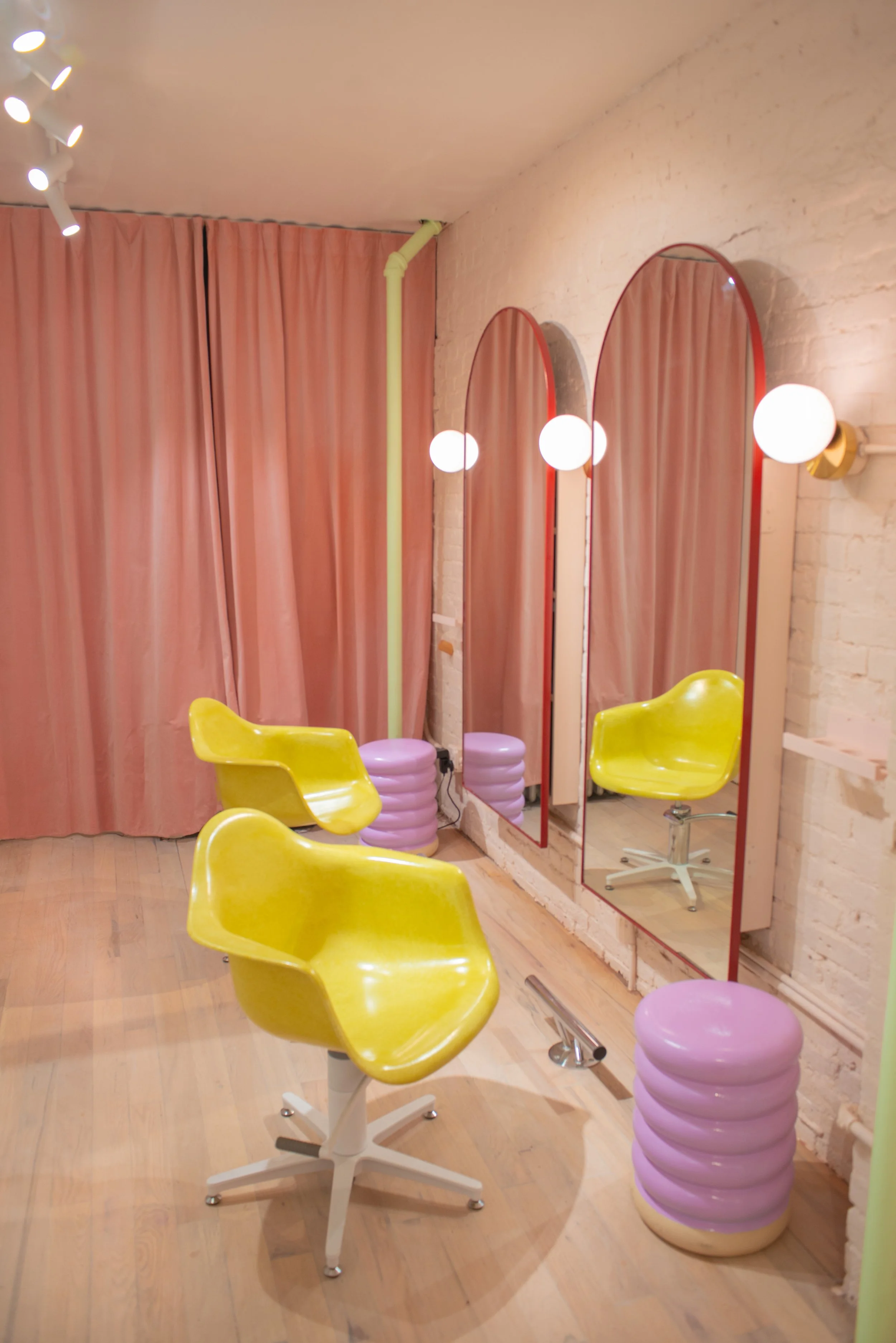 Interior of a hair salon with two large mirrors, yellow salon chairs, pink stool-like footrests, and pink curtains. The lighting includes round bulbs attached to the wall and ceiling.