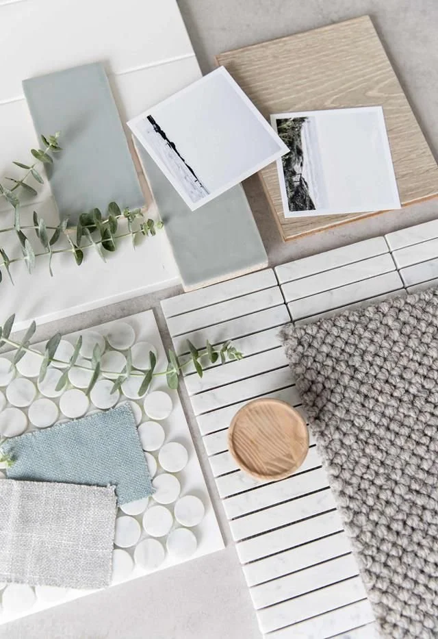A flat lay of interior design materials and swatches, including light-colored tiles, wood samples, fabric swatches, a gray textured rug, and small photographs. There is a sprig of Eucalyptus added for decoration.