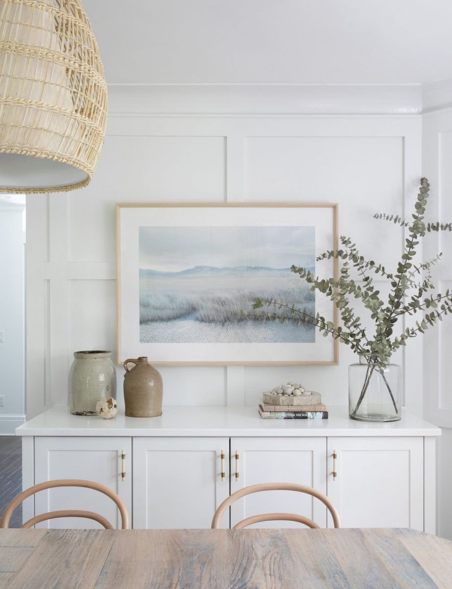 Room interior with wooden dining table, two chairs, a large glass vase with greenery, pottery on white cabinets, framed landscape art, and a wicker pendant light.
