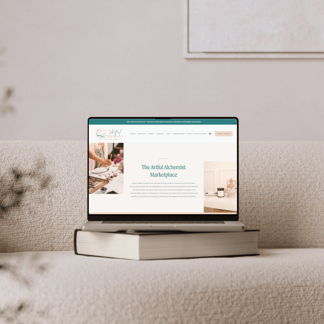 Laptop displaying The Artful Alchemist Marketplace website, resting on a closed book on a beige couch.
