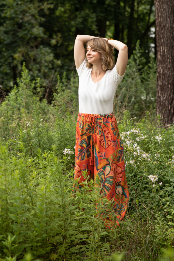 A woman standing in a lush green outdoor setting with her hands behind her head, wearing a white top and colorful patterned wide-leg pants.