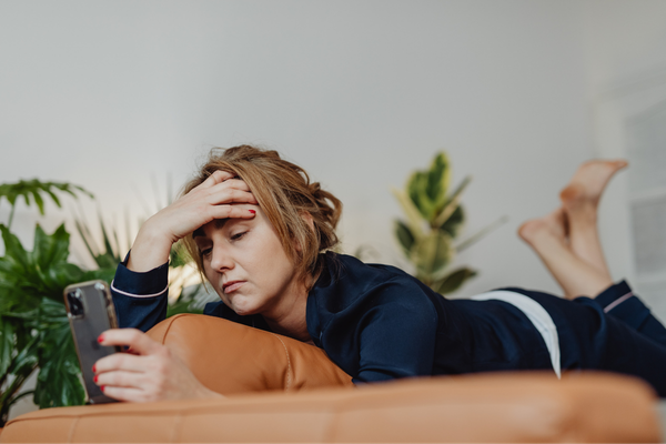 Woman lying on a couch, holding her head with one hand and looking at her phone, appearing stressed or worried.