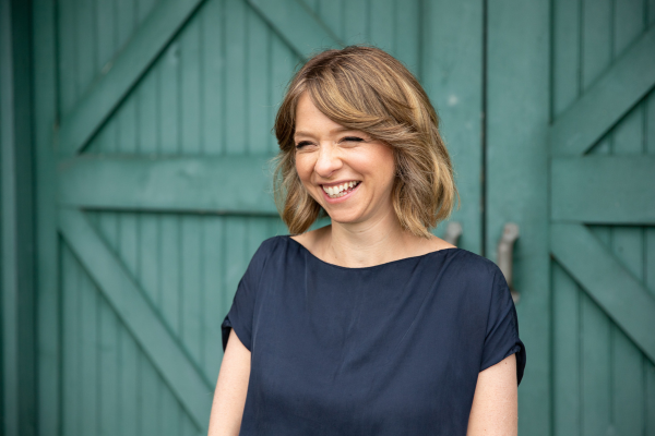 A woman with shoulder-length blonde hair, smiling, wearing a navy blue top, standing in front of green wooden barn doors.