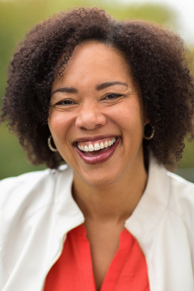 A woman with curly hair smiling outdoors, wearing hoop earrings and a white jacket over a red top.