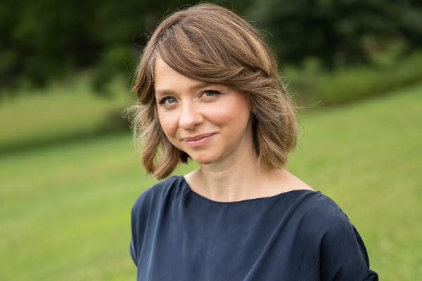 Portrait of a woman with shoulder-length light brown hair, wearing a navy blue top, standing outdoors with a blurred green background.
