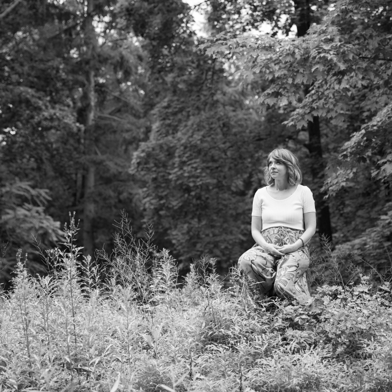 A woman with shoulder-length hair sitting outdoors in a natural setting with trees and plants in black and white.