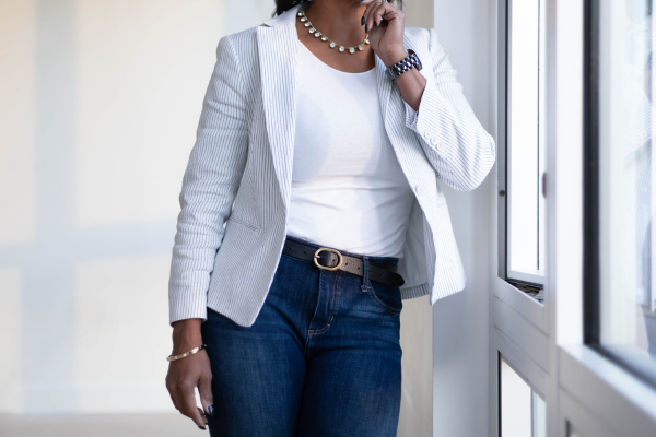 Woman standing near a window, wearing a light-colored blazer, white shirt, jeans, and jewelry.