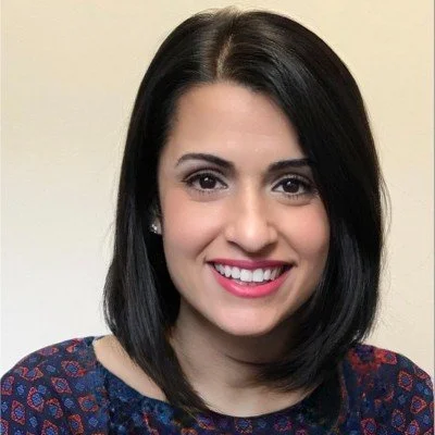 Close-up of a woman with shoulder-length dark hair, smiling, wearing a navy blue top with a red pattern, against a plain light-colored background.