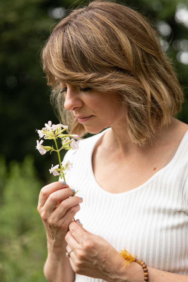 A woman with shoulder-length brown hair, wearing a white ribbed shirt, holding and smelling a light pink flower outdoors.