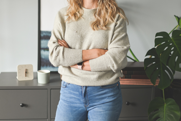 A woman standing indoors with her arms crossed, wearing a beige sweater and blue jeans, next to a plant and a dresser.
