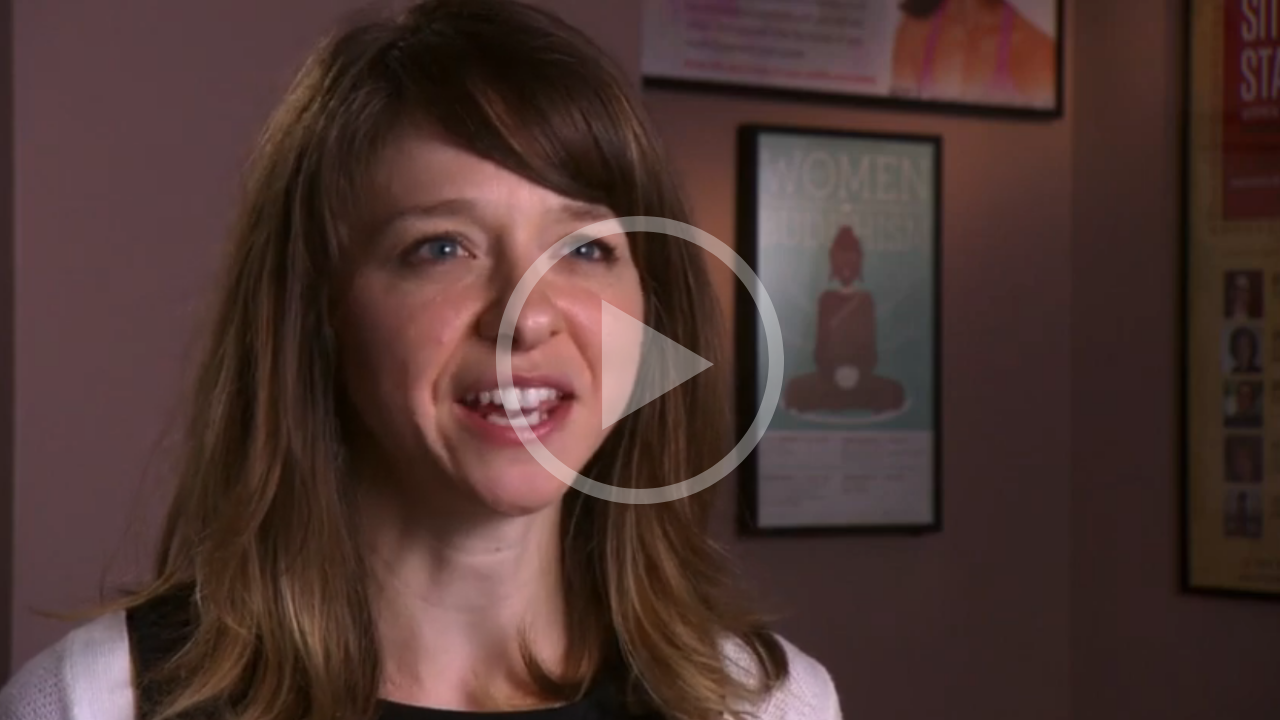 A woman with shoulder-length brown hair and light skin, speaking in an interview setting, with posters on a wall behind her.