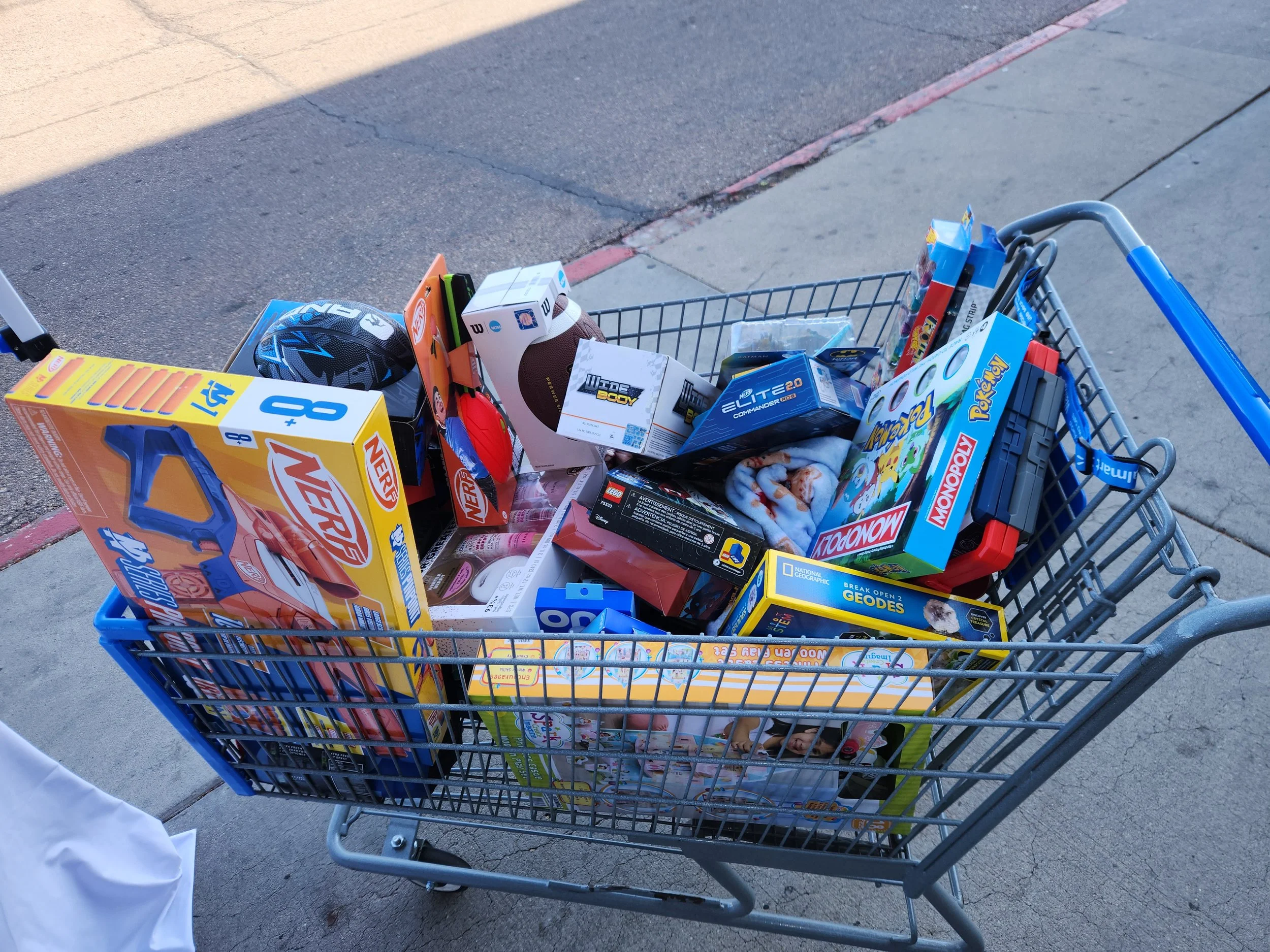A shopping cart filled with toys, including a Nerf blaster, a football, a monopoly game, a puzzle, and other children's toys, is parked on a concrete sidewalk at an outdoor shopping area.