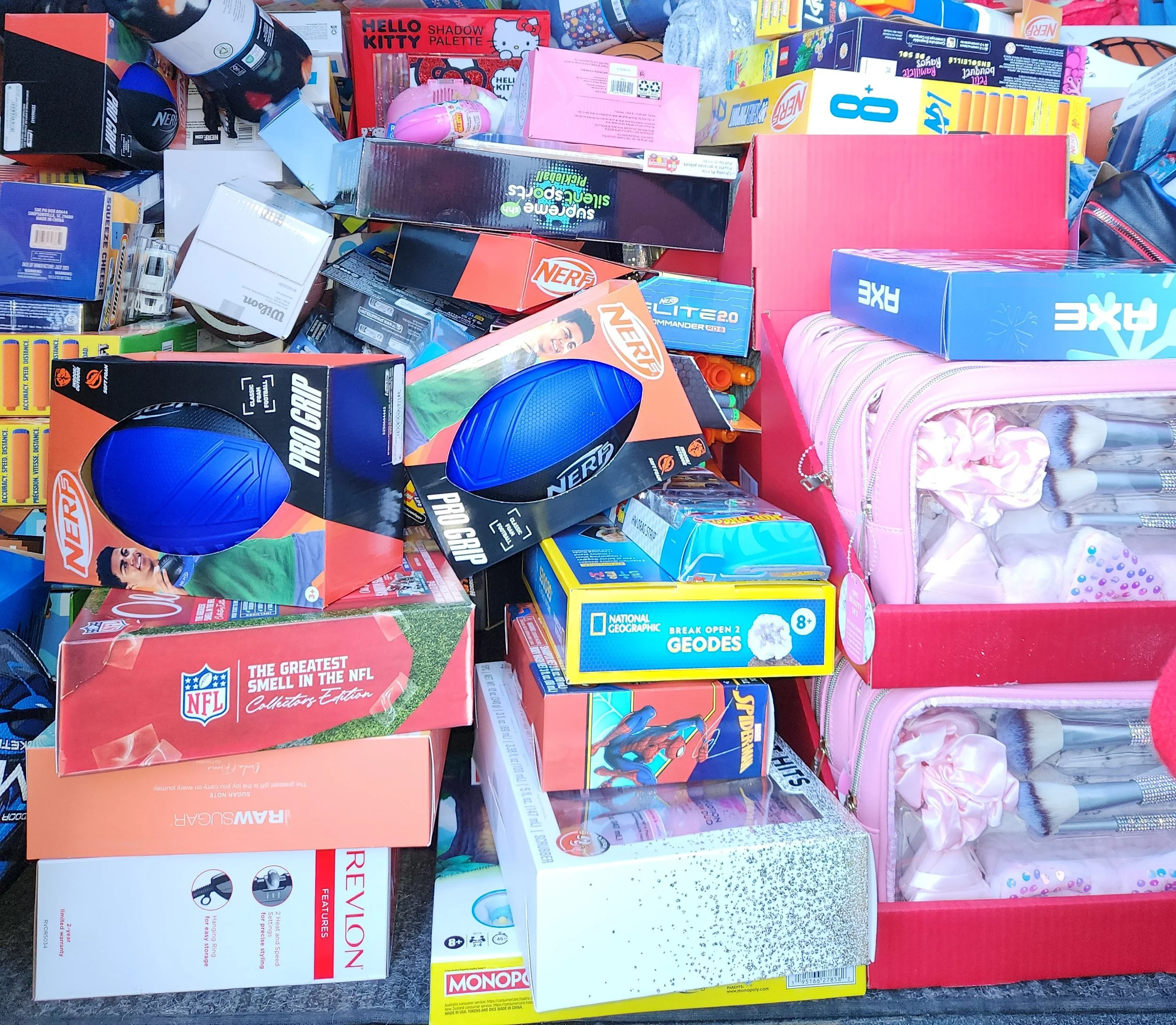 A variety of children's toys, games, and activity sets laid out on a surface, including boxes for Nerf, Monopoly, and National Geographic geodes, as well as plush toys, a pink backpack, and other colorful items.
