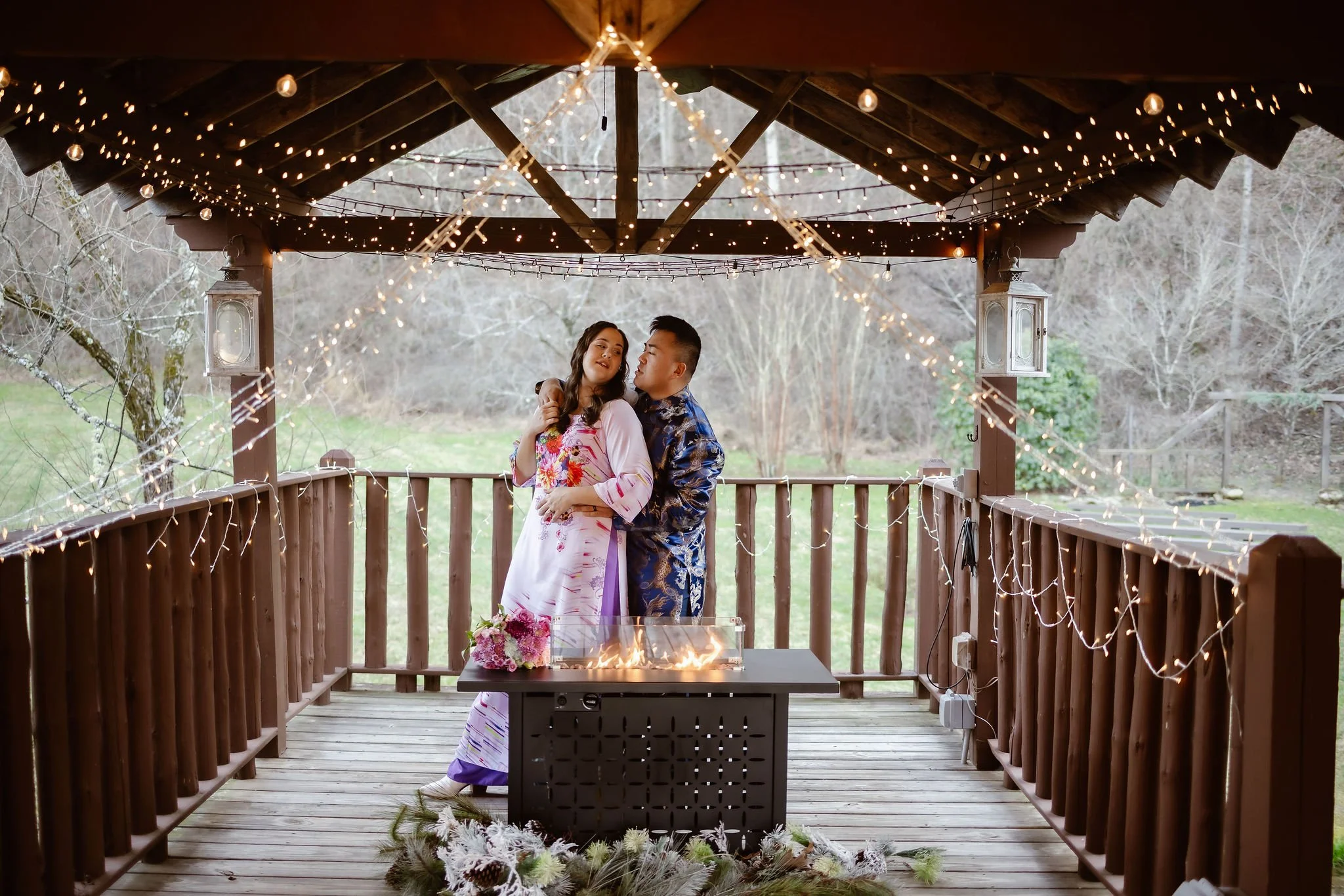 Couple in traditional Vietnamese wedding attire embracing beneath glowing string lights on a rustic pavilion during wedding traditions, photographed by a Gatlinburg photographer while eloping to Gatlinburg.