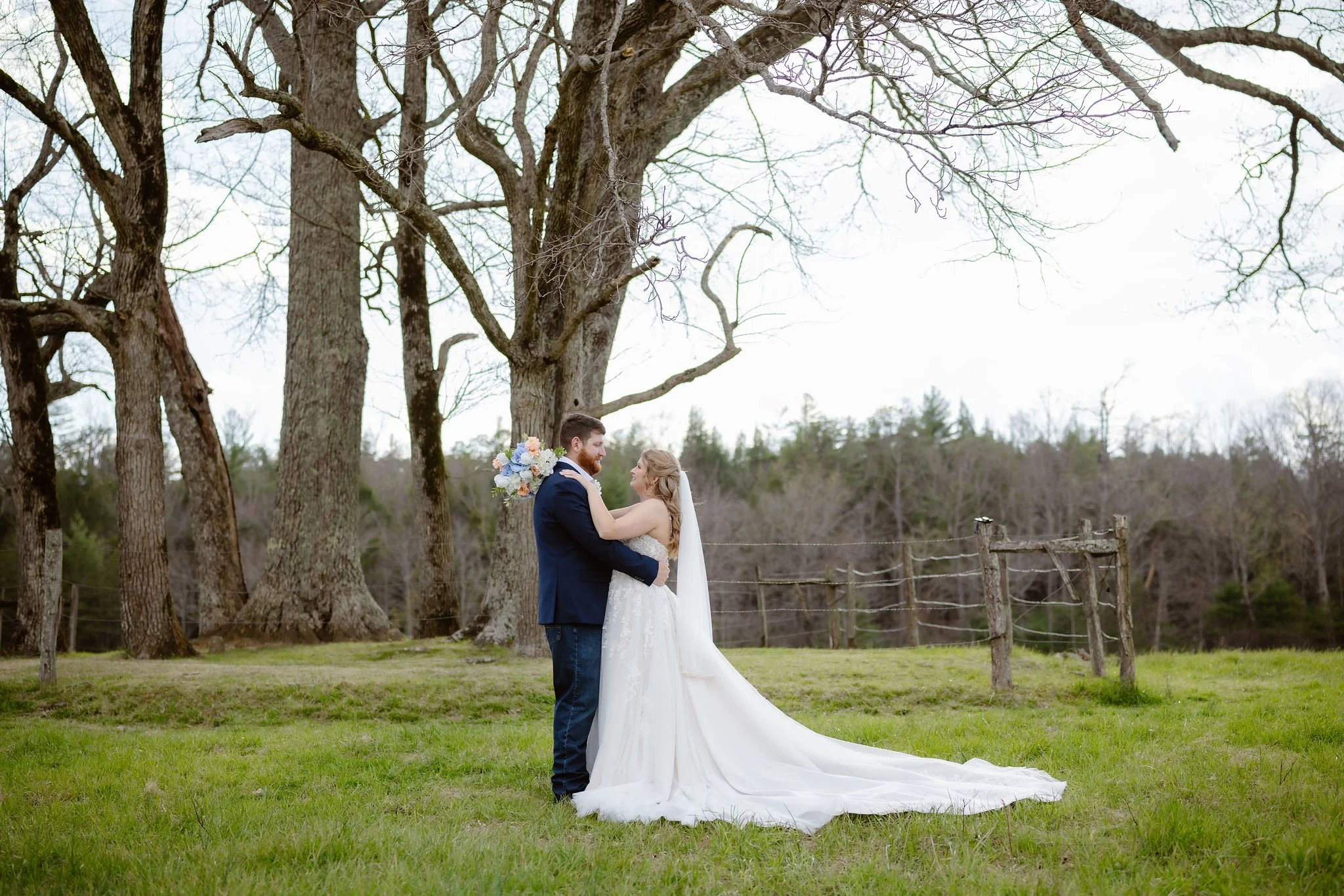 A bride and groom embrace beneath large trees in a grassy field as the bride's wedding dress trails behind her. This outdoor wedding portrait captures the beauty of eloping to Gatlinburg with natural scenery photographed by a Gatlinburg photographer.