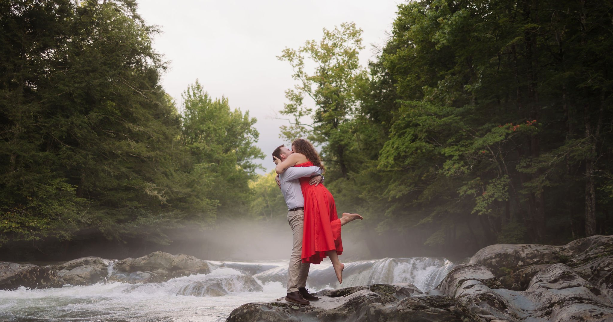 Couple kisses while he lifts her above the rocks beside a misty gatlinburg waterfall in greenbrier.