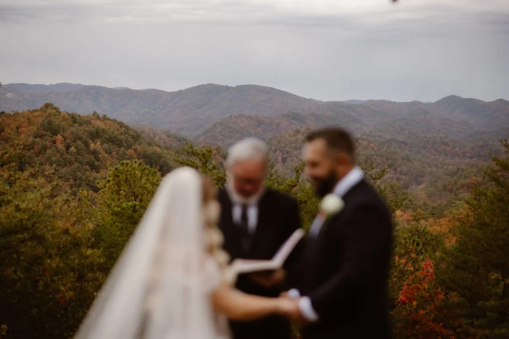 A blurred, artistic perspective of a Foothills Parkway elopement ceremony with the majestic Smoky Mountains in crisp focus behind the bride, groom, and officiant