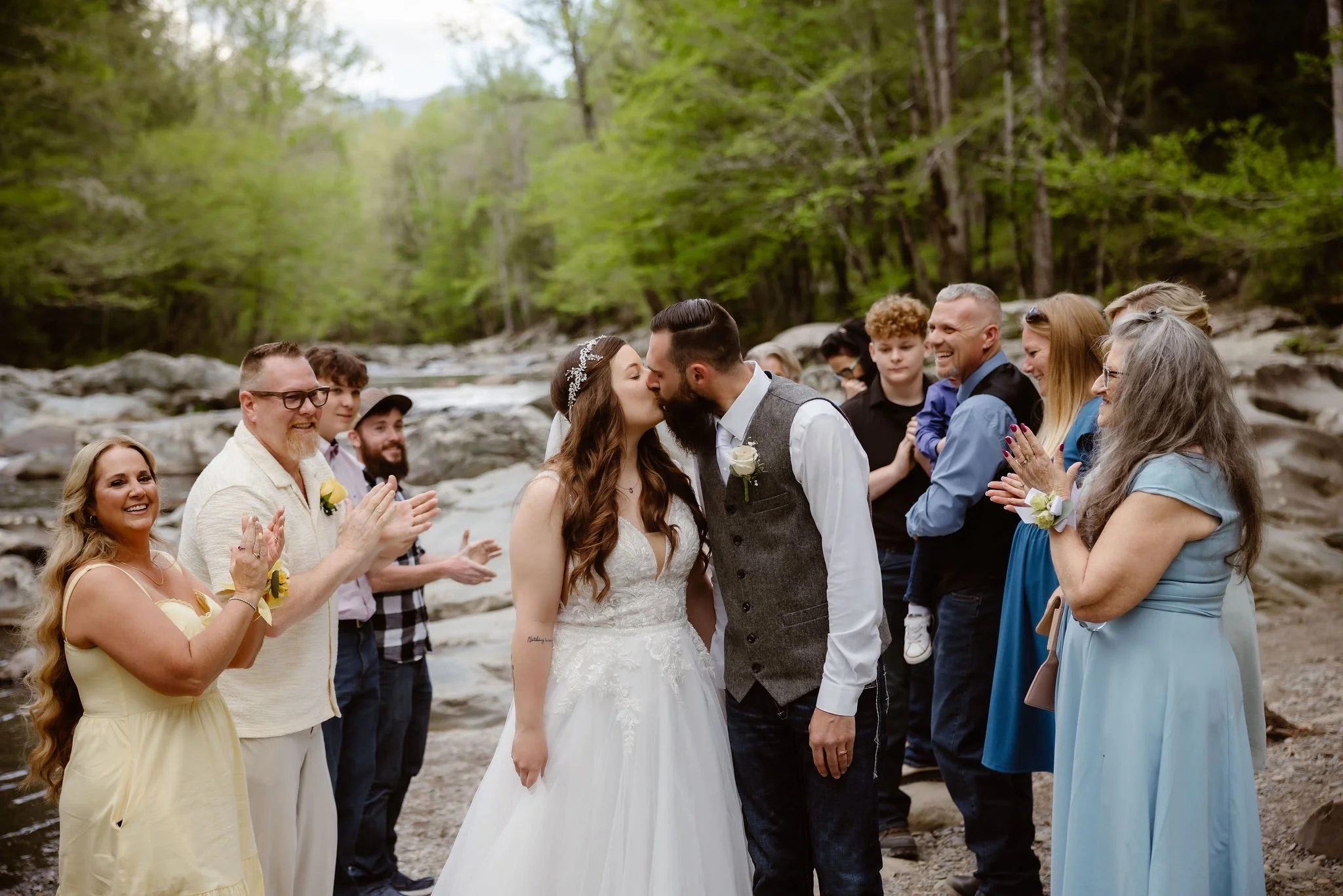 Newlyweds kiss as family and friends applaud beside the river after their smoky mountain elopement ceremony in greenbrier.