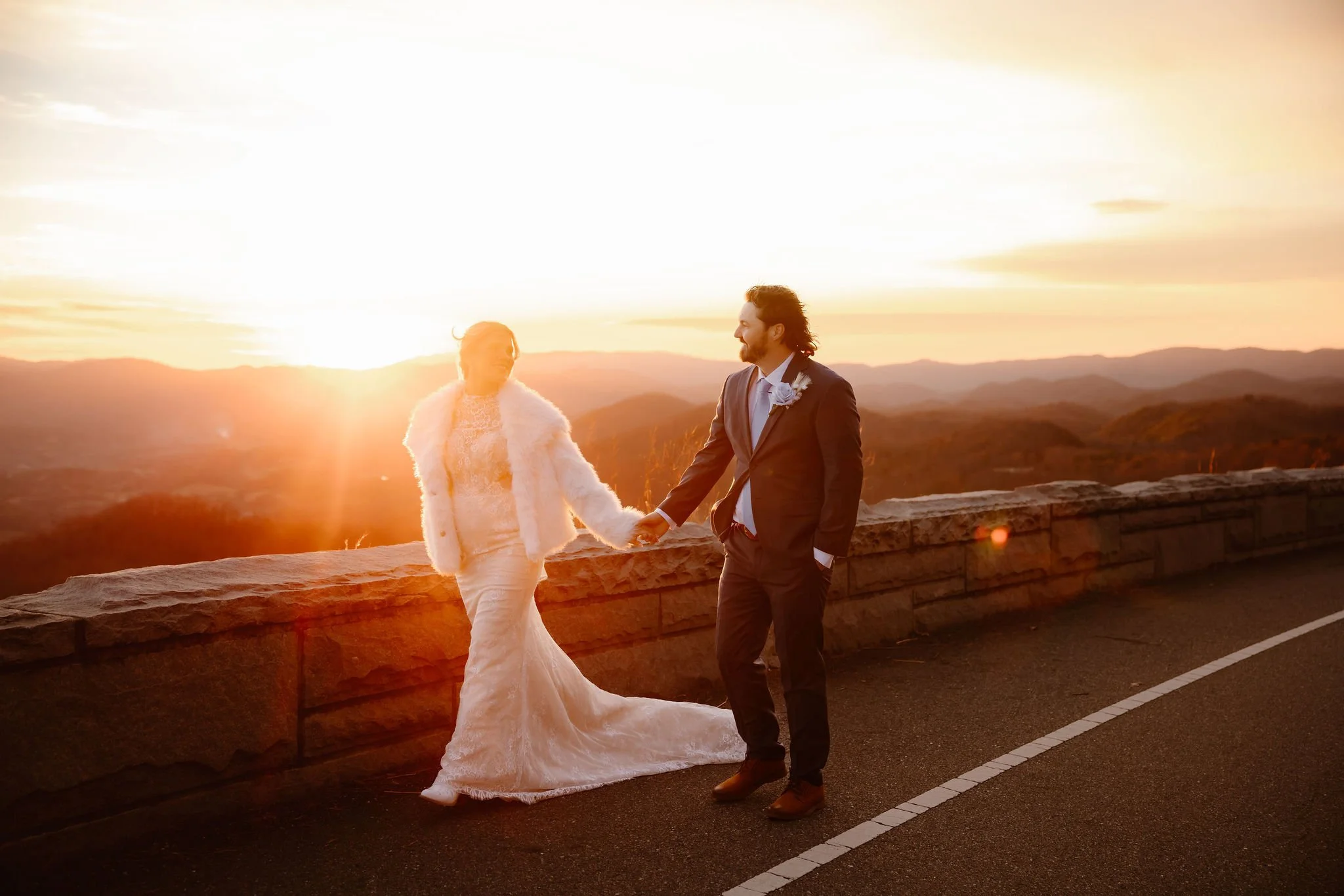 Bride and groom walking hand in hand along Foothills Parkway at golden hour, bathed in the warm glow of the setting sun with a panoramic mountain view behind them. Captured by National Park Photographers in the Smoky Mountains.