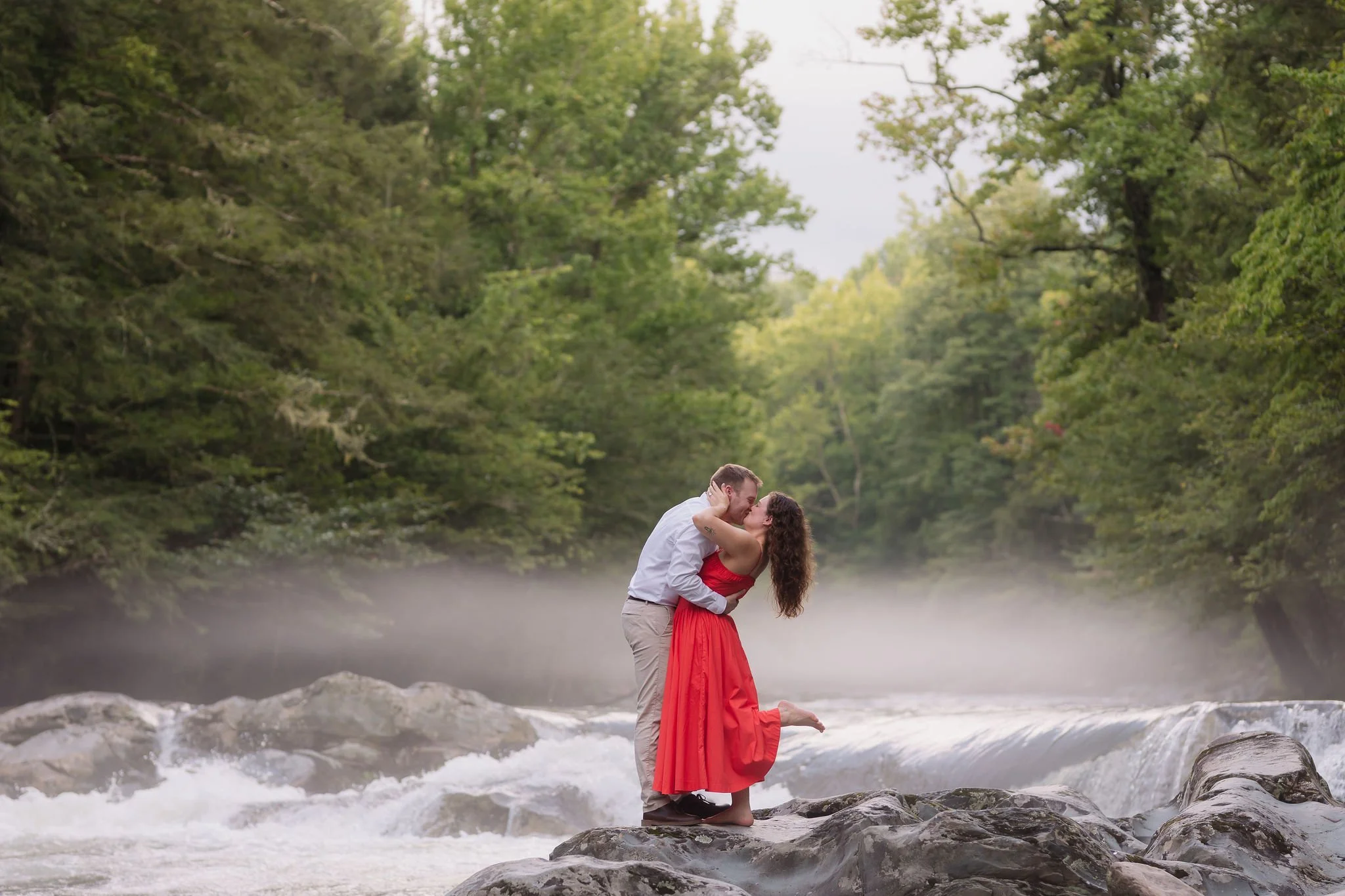 Couple kisses on rocks beside a misty gatlinburg waterfall in greenbrier during their outdoor engagement session.