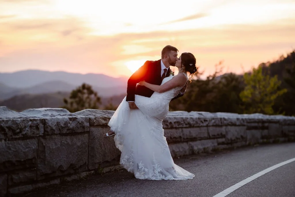 A groom dips his bride for a kiss as the sun sets over the Smoky Mountains along the Foothills Parkway