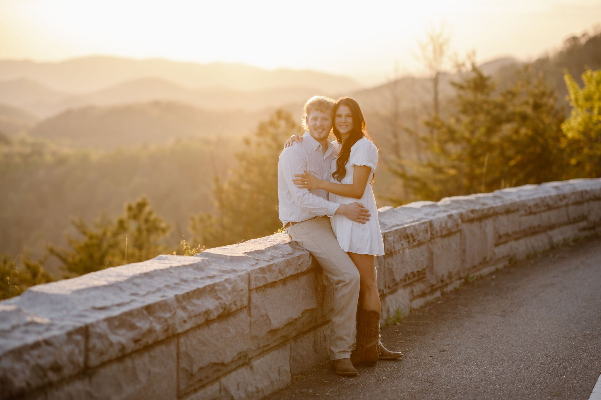 Couple embracing at a scenic mountain overlook during golden hour, smiling at the camera with layered mountain views and a stone overlook wall behind them