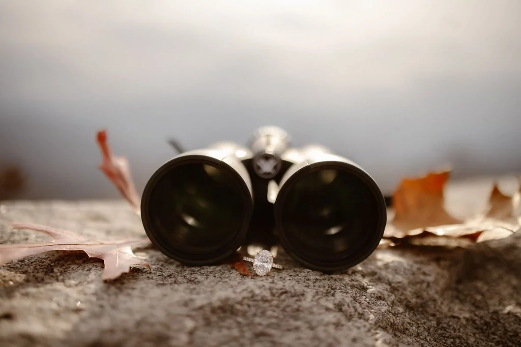 Close-up of engagement ring placed in front of binoculars on stone overlook at Foothills Parkway, styled engagement photos clothes details captured by Gatlinburg photographer during creative mountain proposal session