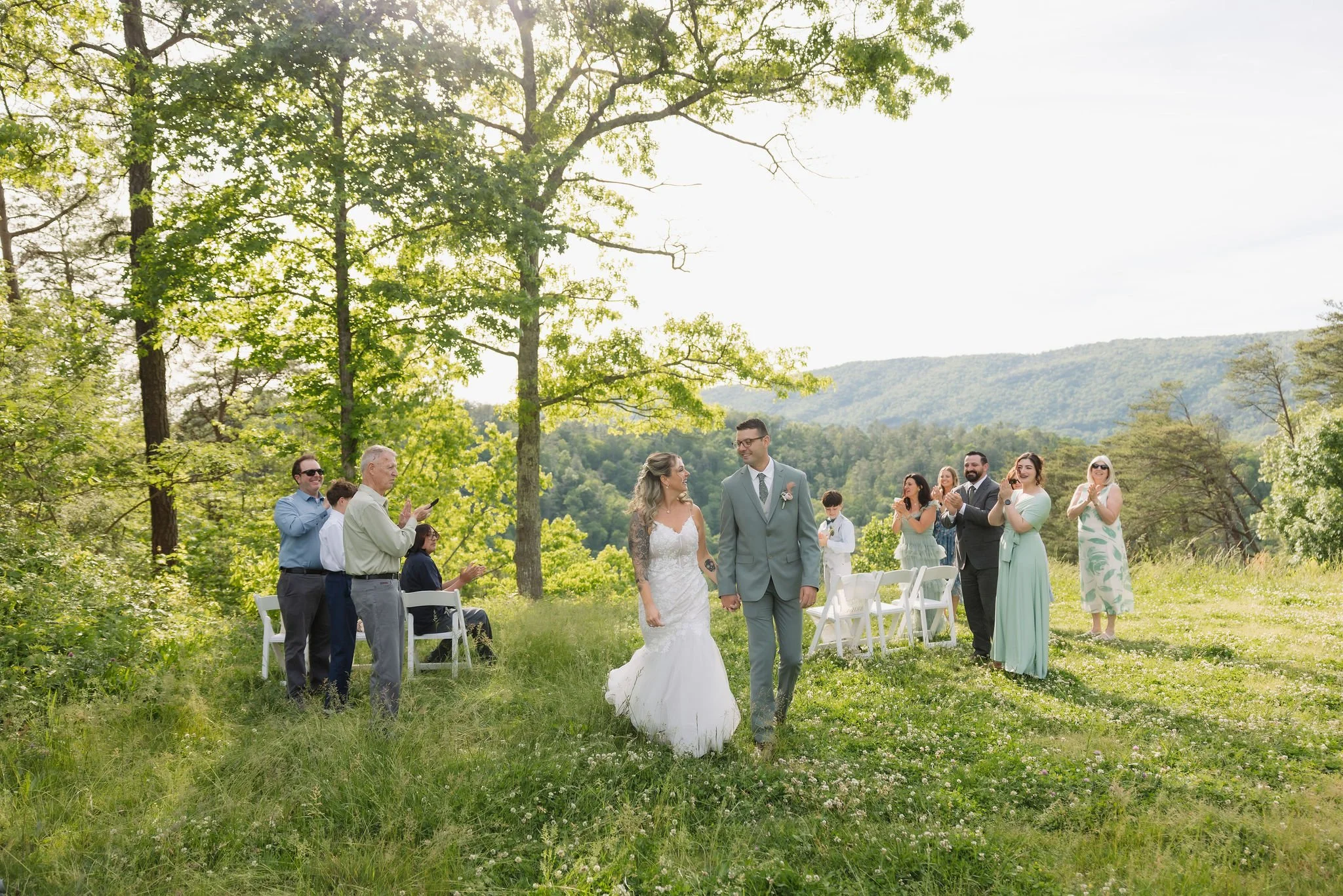 Bride and groom walk hand in hand through a sunlit meadow as guests applaud after their Smoky Mountain Ceremony overlooking the mountains.
