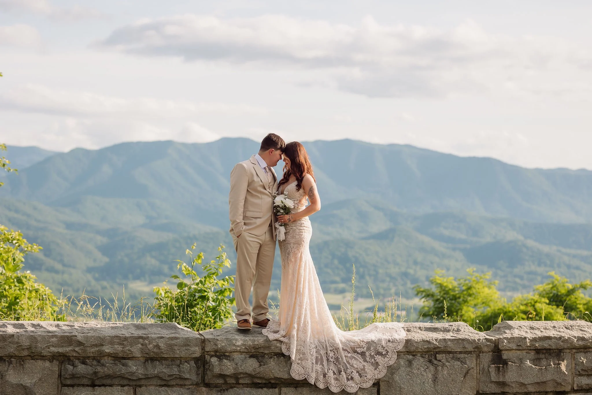 A bride and groom touch foreheads at a scenic Foothills Parkway overlook, her intricate lace wedding train draped over the stone wall with sweeping Smoky Mountain views behind them.