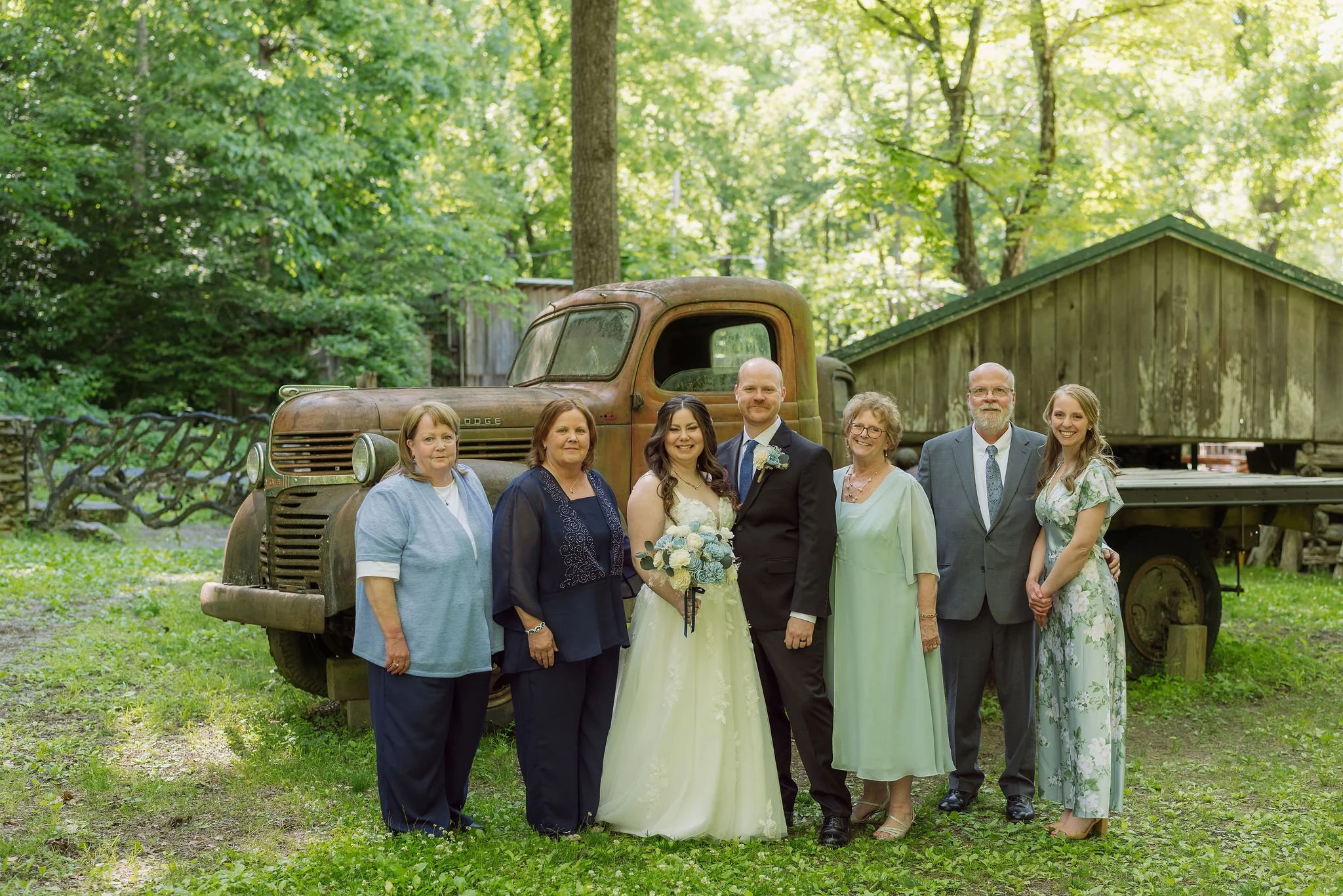 Newlyweds pose with family beside a vintage truck and rustic buildings after their Greenbrier wedding ceremony in the Smoky Mountains.