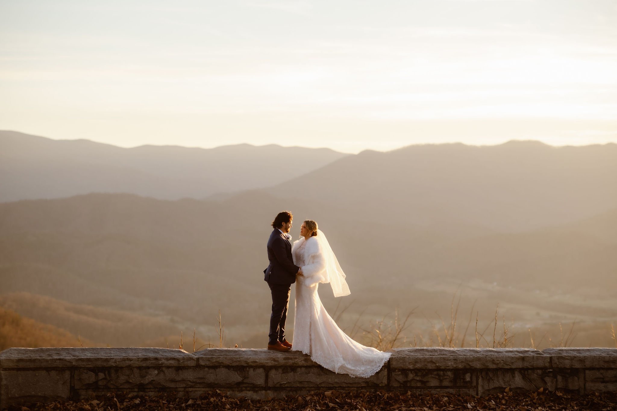 Bride and groom standing on a stone overlook at Foothills Parkway, facing each other with a sweeping view of the Smoky Mountains behind them at sunset, captured by National Park Photographers.