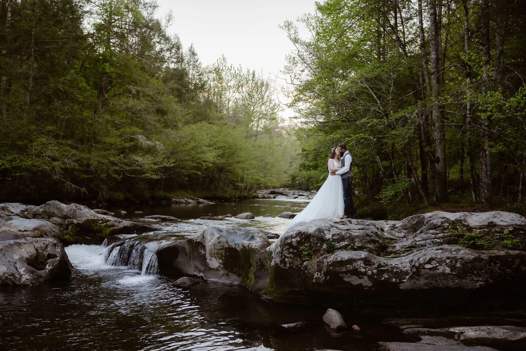 Wide portrait of newlyweds kissing beside a rocky river and small cascades surrounded by the dense forest of greenbrier.