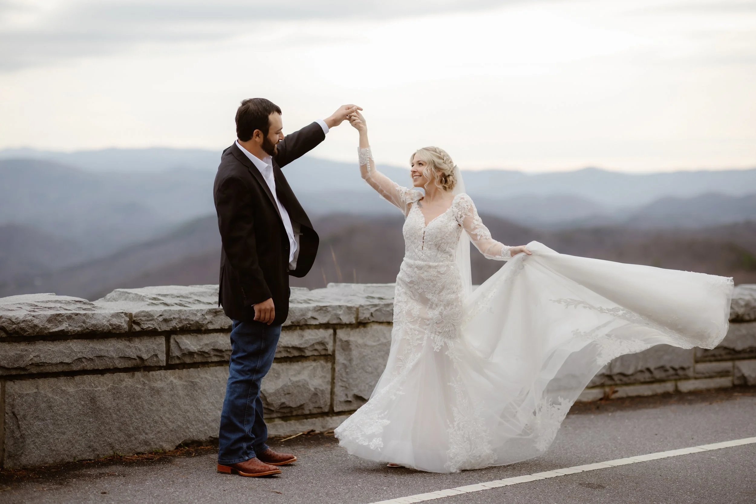 Bride twirling in her lace wedding dress while holding hands with her groom at a scenic mountain overlook during a Smoky Mountain elopement near Parkside resort, photographed by a Gatlinburg photographer under cloudy skies.