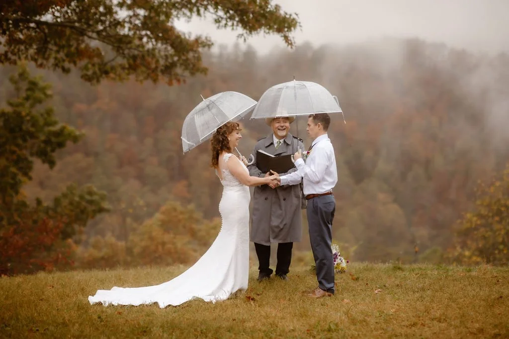 Couple exchanging vows under clear umbrellas during rain on my wedding day Foothills Parkway elopement, captured by Gatlinburg photographer with foggy mountain views, autumn foliage, and intimate rainy outdoor wedding ceremony
