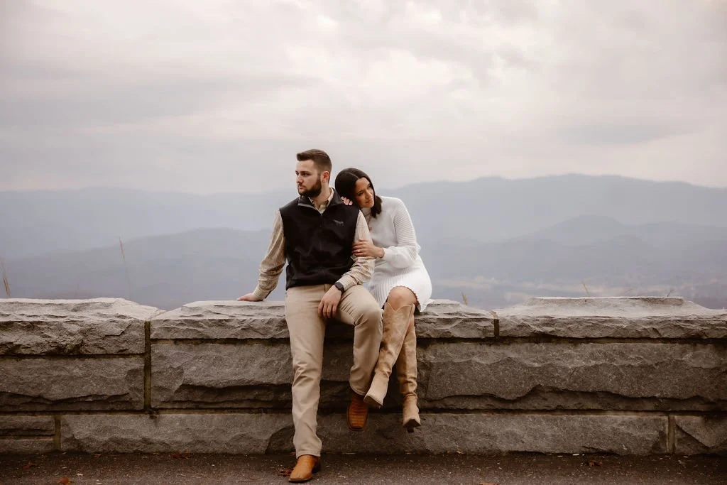 Couple sitting on stone overlook wall in coordinated neutral outfits with mountain view, Gatlinburg photographer capturing stylish fall engagement session with engagement photos clothes inspiration in the Smoky Mountains