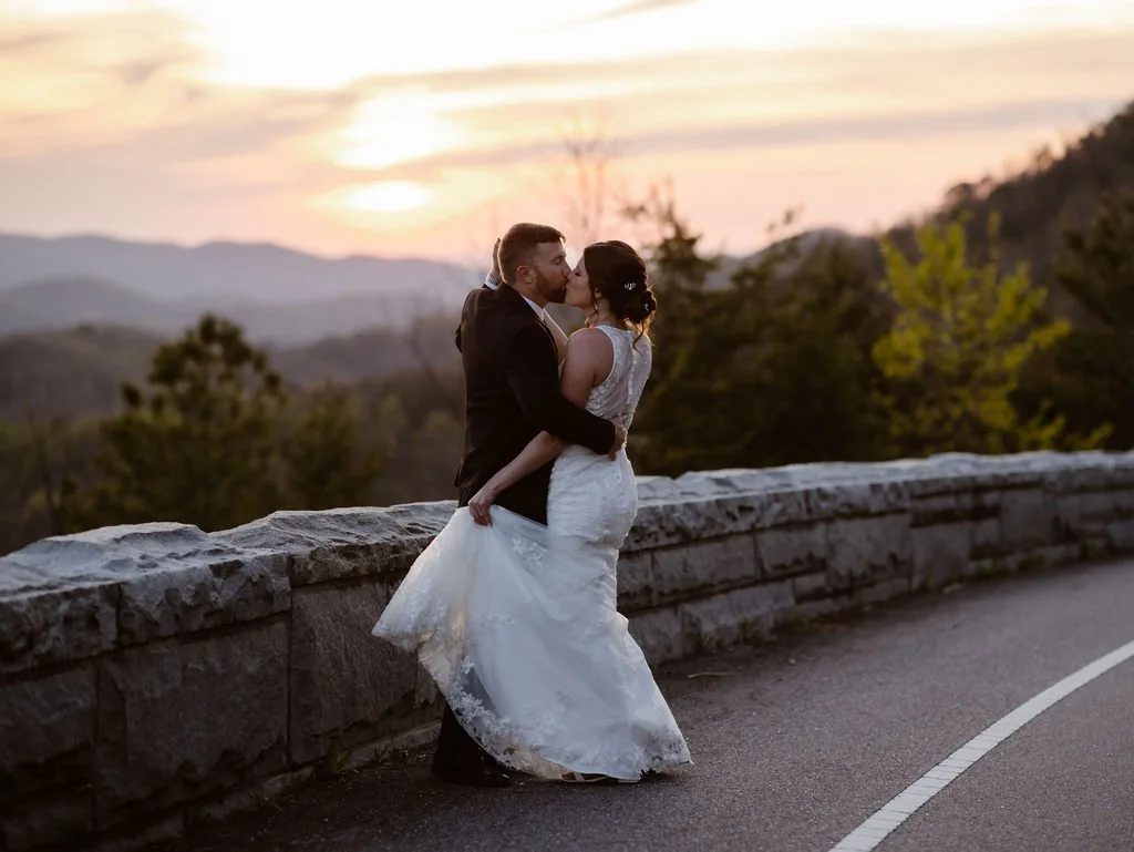 A bride and groom share a kiss at sunset along the Foothills Parkway, with the Smoky Mountains stretching across the horizon behind them