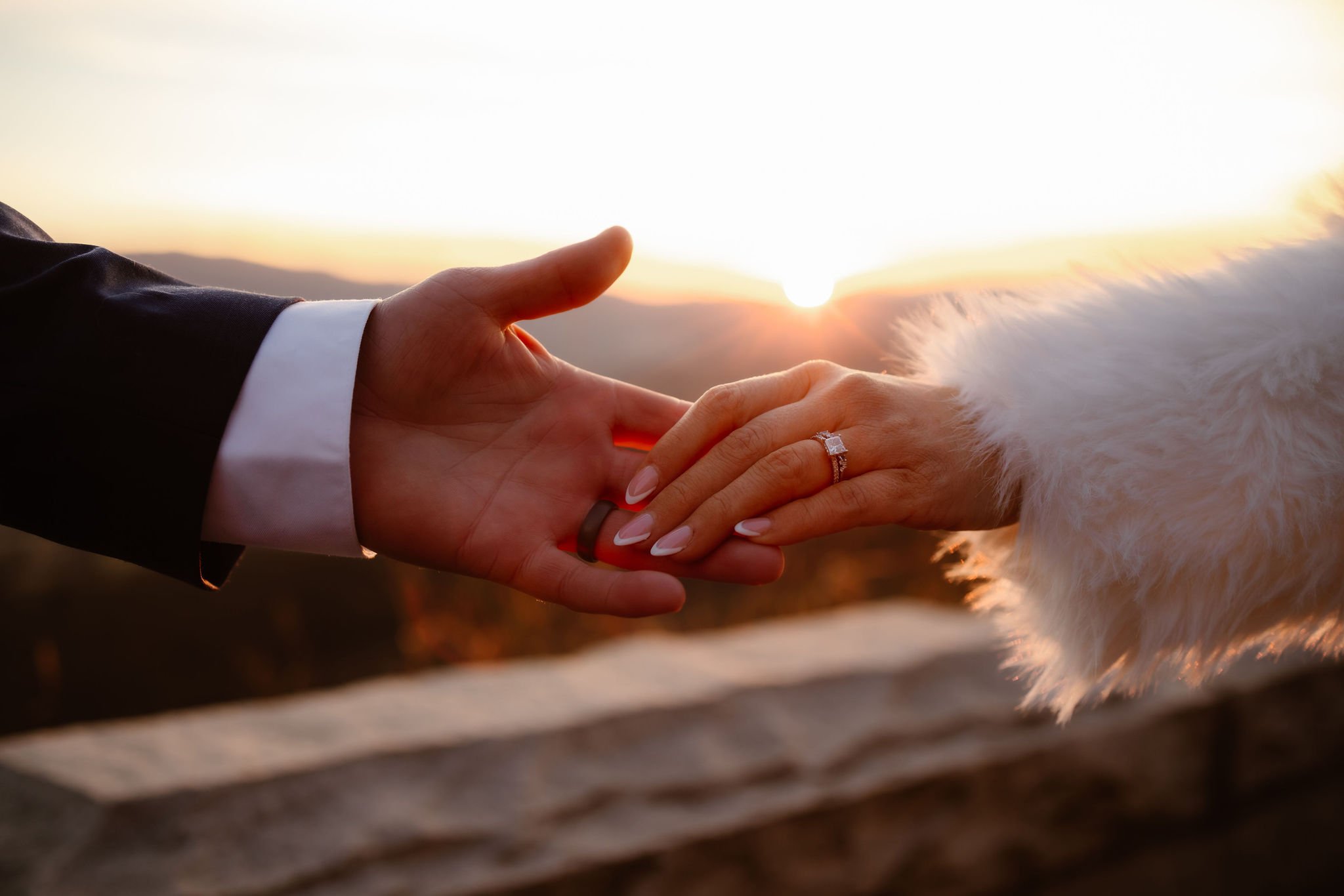 Close-up of the bride and groom’s hands reaching toward each other at sunset, highlighting the bride’s engagement ring and faux fur sleeve, with a mountain view along Foothills Parkway in the background. Captured by National Park Photographers.