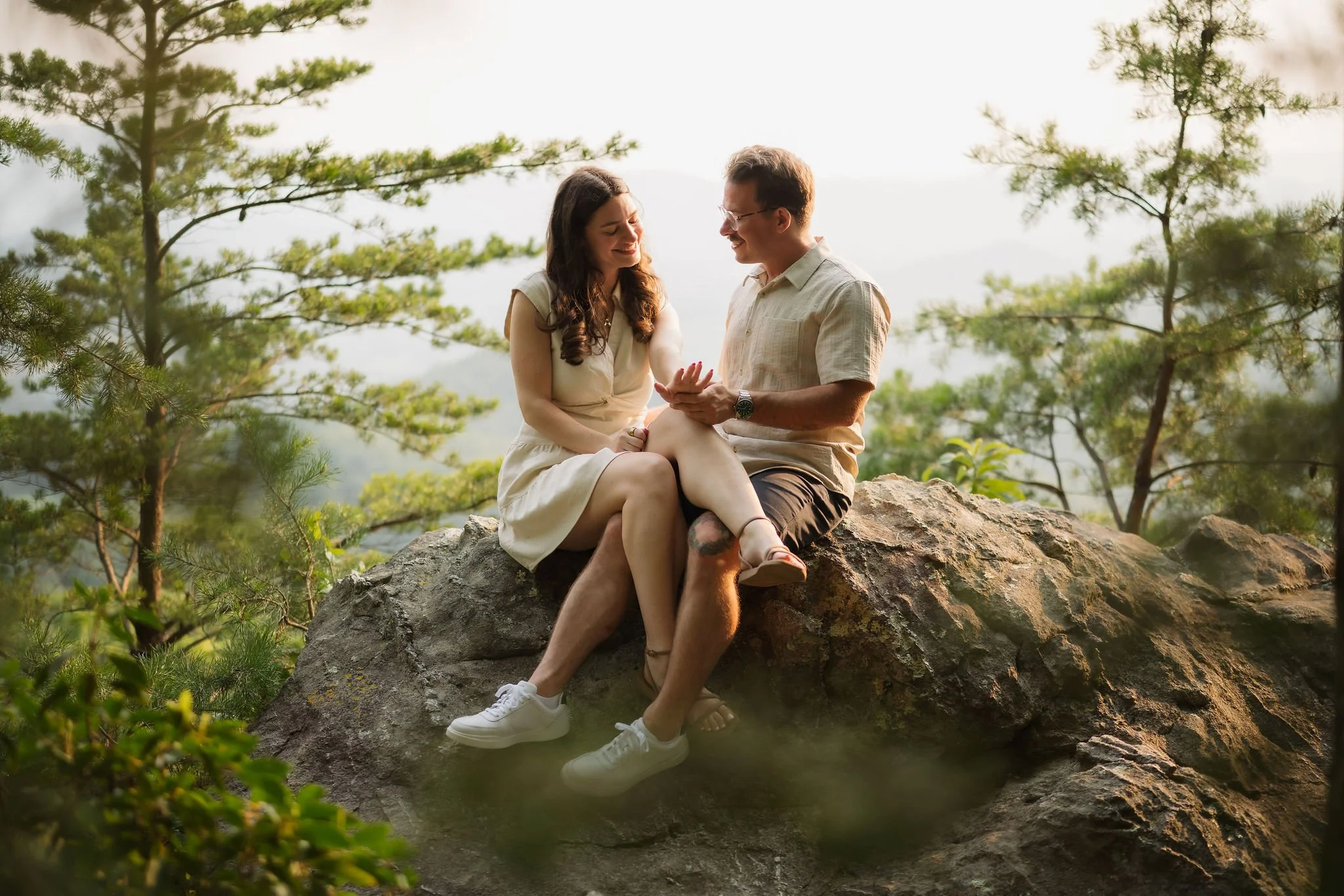 Smoky Mountain engagement session featuring a couple sitting together on a large rock overlook, admiring an engagement ring with mountain views and pine trees in the background, captured by a Gatlinburg photographer.