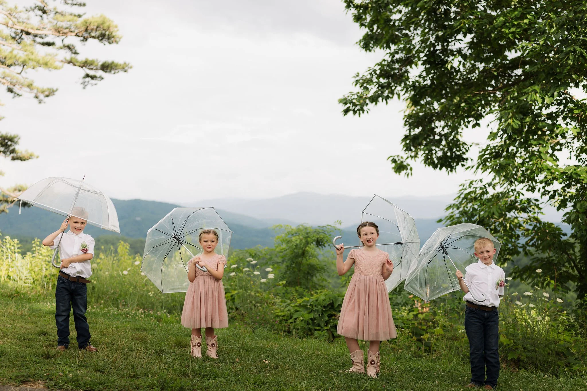 Four children in blush pink dresses, cowboy boots, and white shirts play with clear umbrellas against the misty Smoky Mountain foothills during a spring Gatlinburg elopement.
