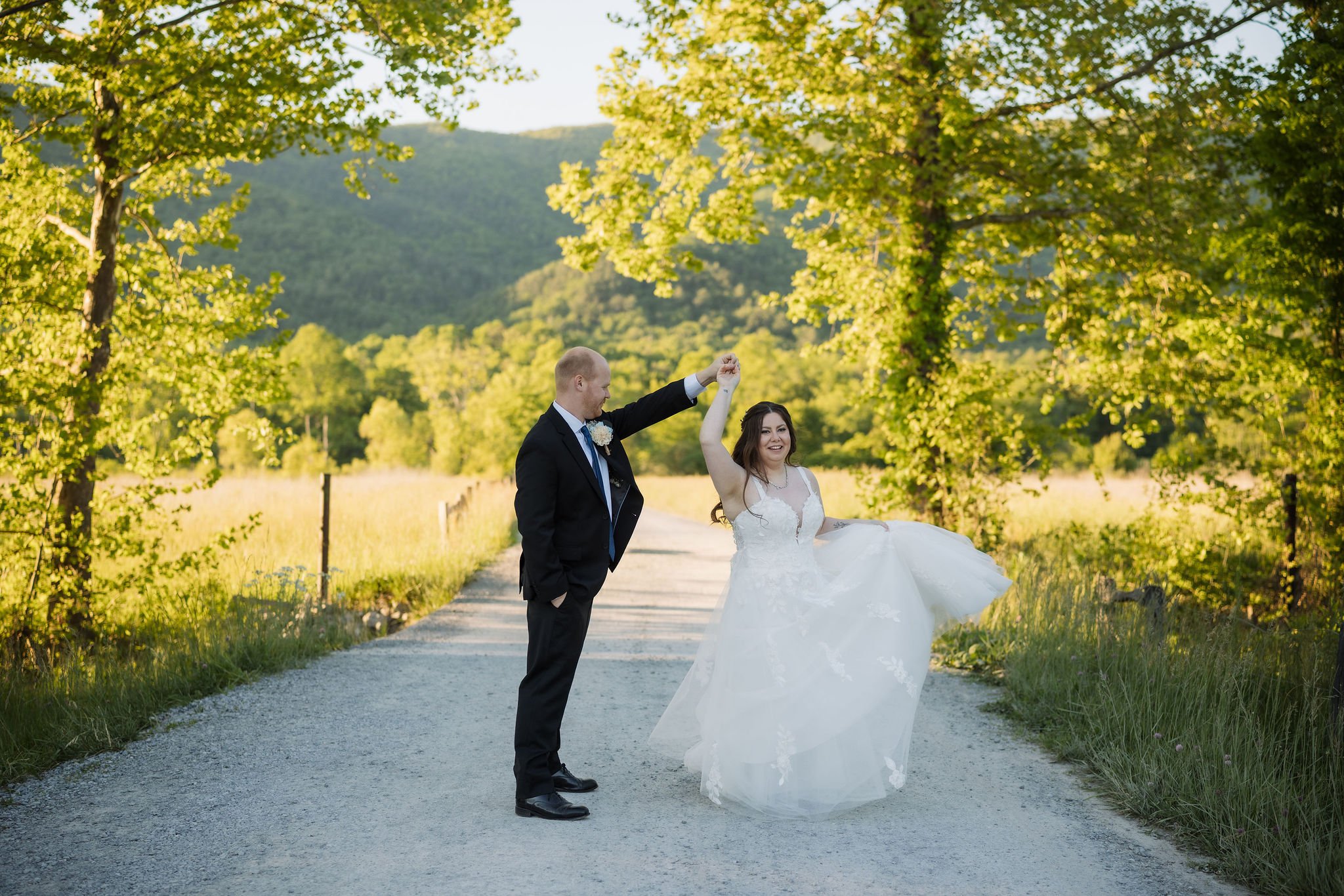 Bride twirls in her wedding dress as the groom holds her hand on a quiet country road surrounded by mountain views and summer greenery in the Smoky Mountains, captured by a Gatlinburg wedding photographer.
