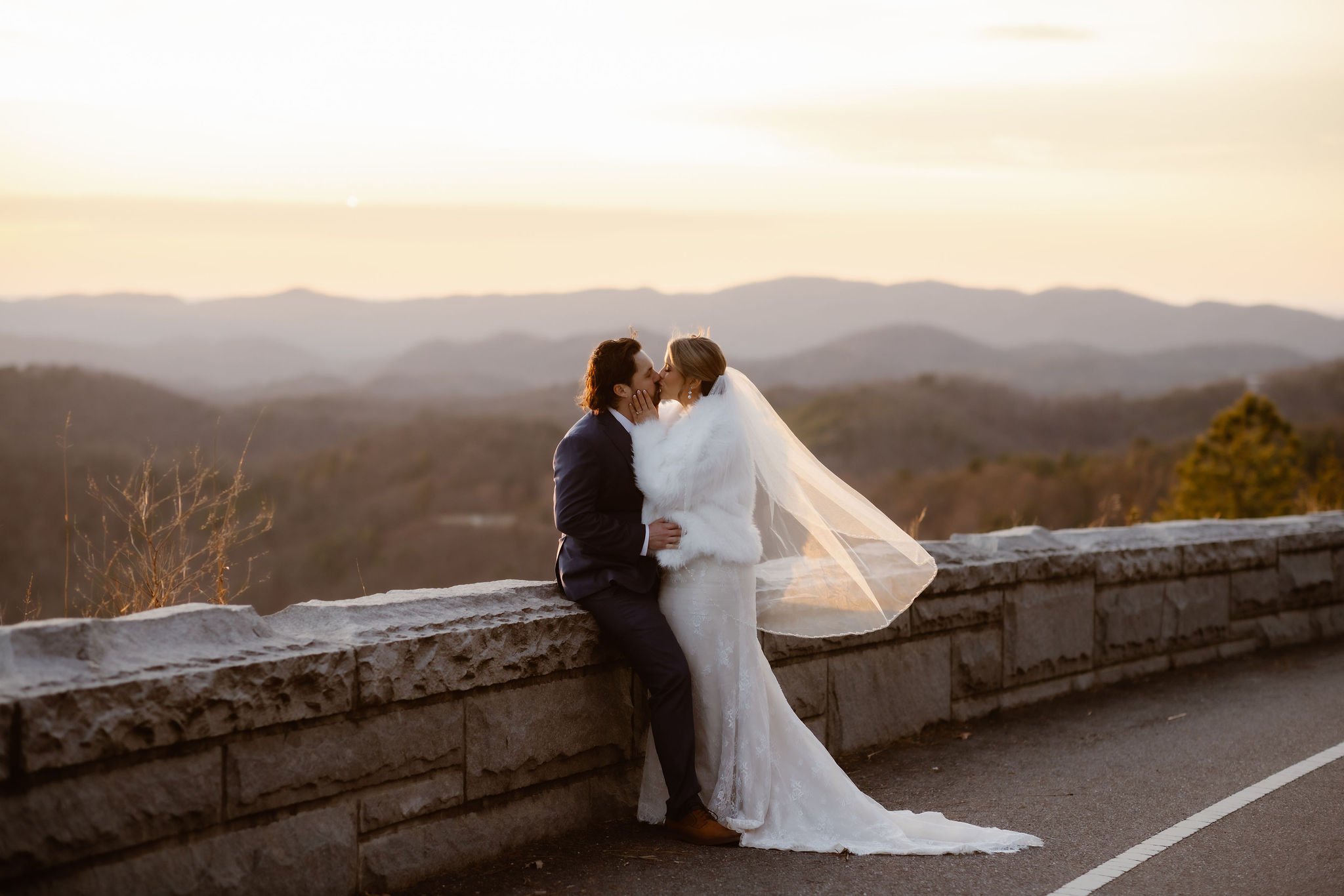 Bride and groom sharing a kiss on a stone ledge at sunset with panoramic mountain views behind them, captured by National Park Photographers along Foothills Parkway in the Smoky Mountains