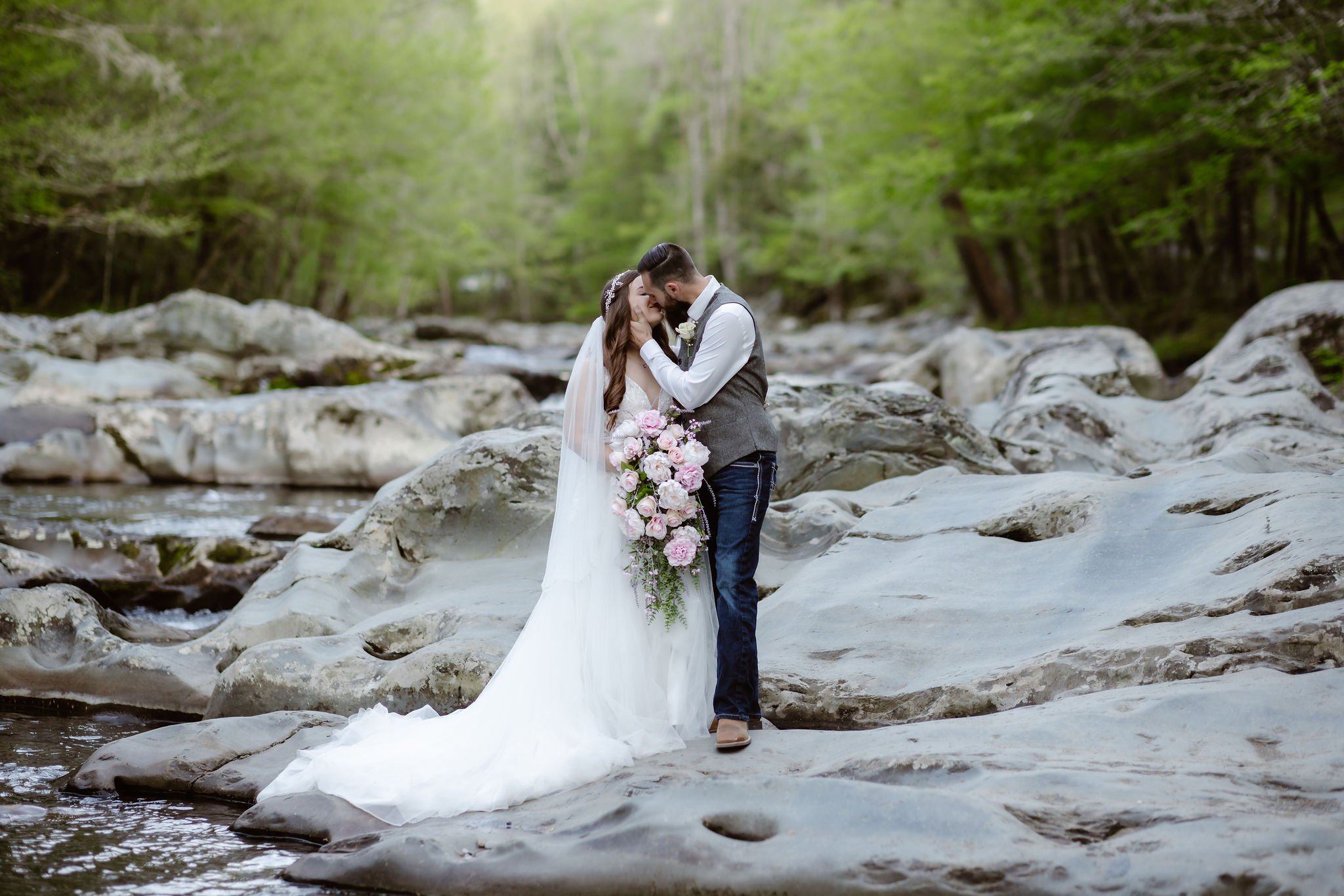 A bride and groom share a kiss on the smooth river rocks during elopement photography at Greenbrier in the Smoky Mountains, captured by a Gatlinburg wedding photographer.