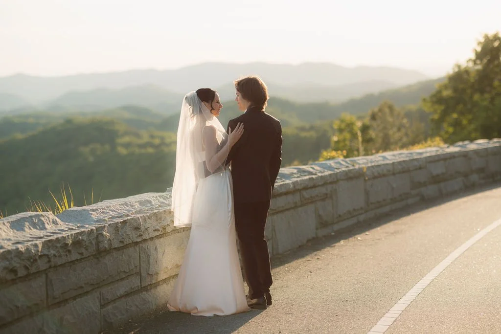 Bride and groom stand together beside the foothills parkway at sunset, overlooking the layered green ridges of the Smoky Mountain foothills.