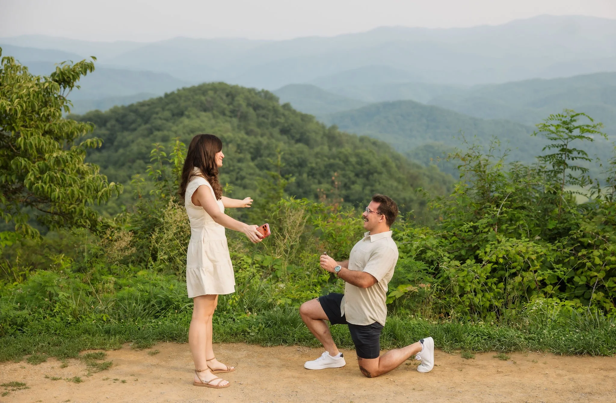 Smoky Mountain engagement at a panoramic mountain overlook as a woman reaches toward her newly engaged partner after a surprise proposal, captured by a Gatlinburg photographer.