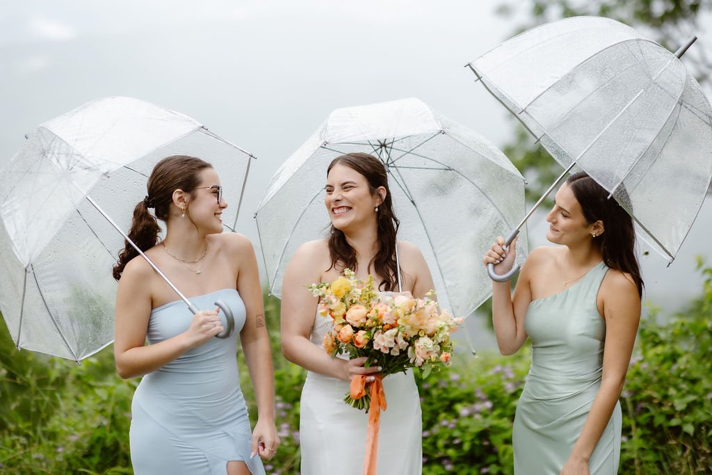 Bride and two bridesmaids standing outdoors under clear umbrellas during a light rain, surrounded by lush greenery and spring blooms, with the bride holding a pastel bouquet