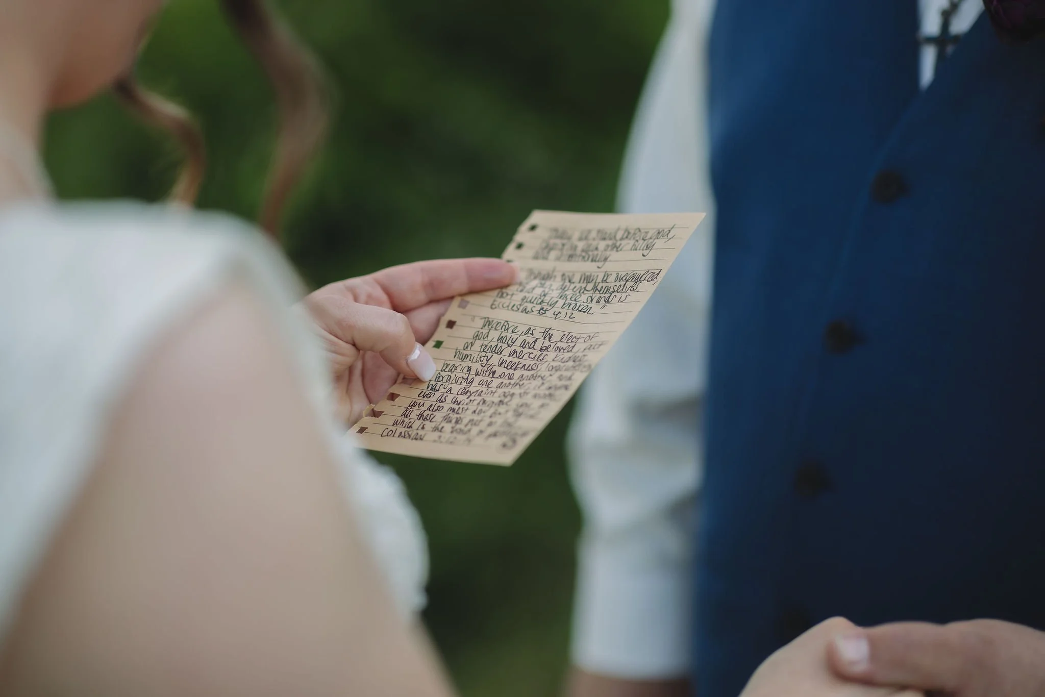 Close-up of a bride holding handwritten wedding vows on lined paper while standing across from her groom in a navy vest during their intimate Gatlinburg elopement ceremony.
